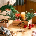 mainile unei femei relalizand o decoratiune intr-un cosulet de craciun, cu globuri, lumanari si crengute de brad