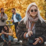 Fată blondă îmbrăcată la modă, cu ochelari de soare și aparat de fotografiat, se distrează cu prietenii ei în parcul public de toamnă.